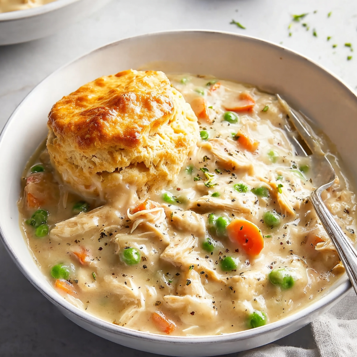 crockpot chicken pot pie A bowl of creamy chicken pot pie stew with shredded chicken, carrots, peas, and onions, topped with a golden biscuit, served on a light-colored table with a fork on the side.