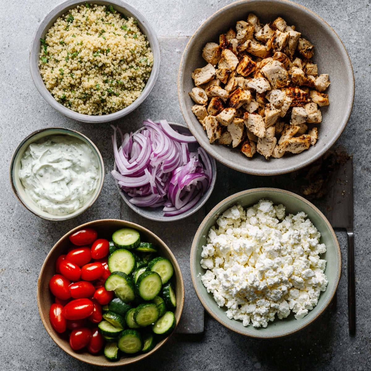 Healthy Greek Bowl Recipe with Chicken & Tzatziki (35 Min) 12 Greek chicken bowl ingredients arranged in bowls including cooked quinoa, grilled chopped chicken, cucumbers, cherry tomatoes, red onion, feta cheese, and tzatziki sauce
