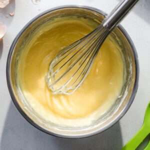 Best Japanese Pancakes Recipe 15 Overhead view of a stainless steel mixing bowl with yellow pancake batter being whisked, metal whisk inside the bowl, green silicone spatulas nearby, cracked eggshells on a light gray kitchen surface, bright natural light, baking preparation scene, realistic food photography, sharp focus, minimal modern kitchen style.