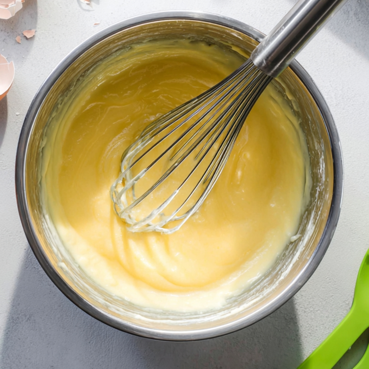 Best Japanese Pancakes Recipe 10 Overhead view of a stainless steel mixing bowl with yellow pancake batter being whisked, metal whisk inside the bowl, green silicone spatulas nearby, cracked eggshells on a light gray kitchen surface, bright natural light, baking preparation scene, realistic food photography, sharp focus, minimal modern kitchen style.