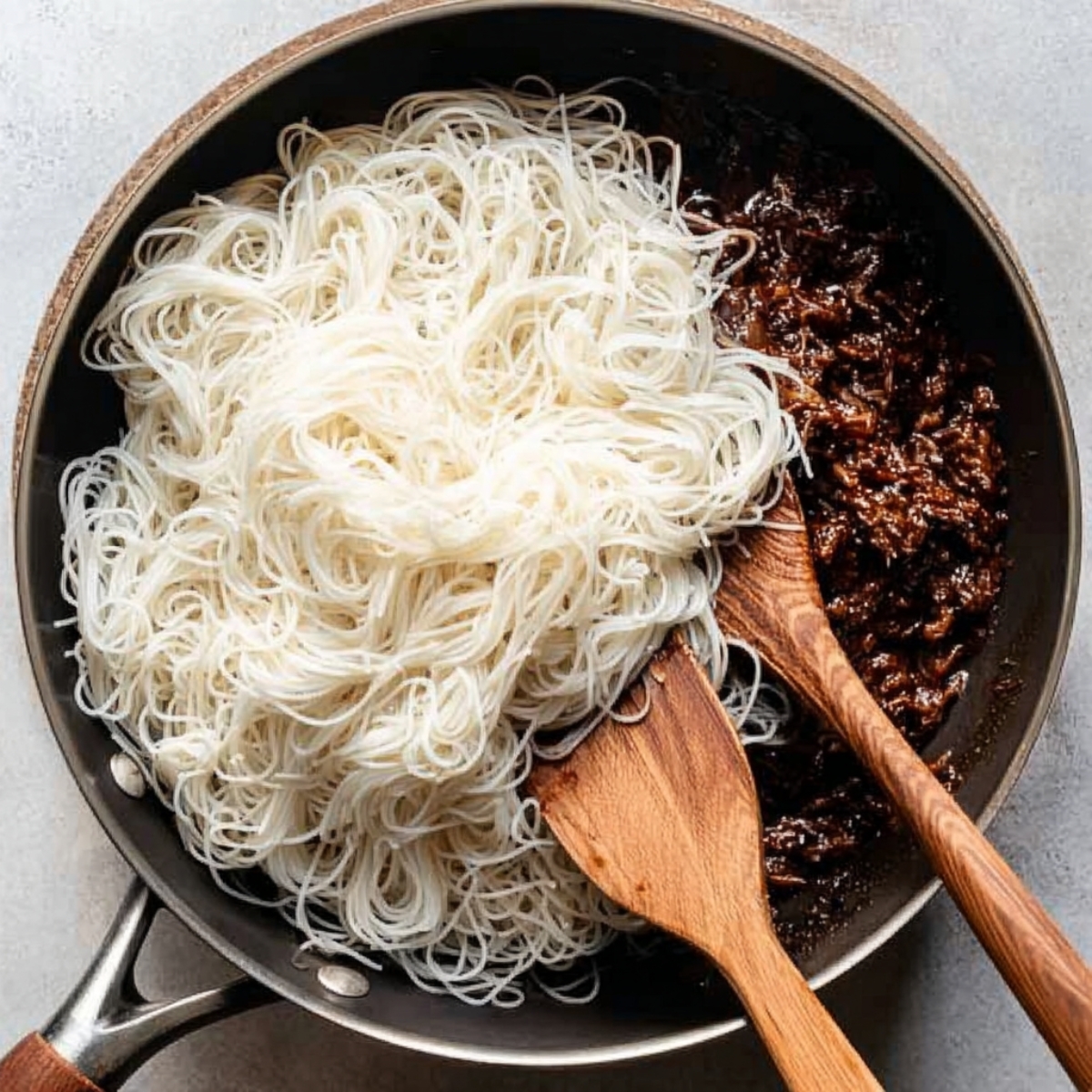 Easy Mongolian Ground Beef Noodles 12 Cooked white rice noodles being added to a skillet with dark ground beef sauce, wooden spoon visible, overhead cooking scene.