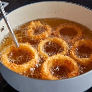 Easy Onion Rings Recipe | 8 Steps to Crispy Perfection 17 Onion rings frying in hot oil inside a white pot, bubbling oil action shot, golden rings floating, cooking thermometer visible, realistic food photography, dynamic kitchen scene.