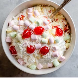 Ambrosia Fruit Salad Recipe 14 “Overhead shot of a white bowl filled with ambrosia salad ingredients including mini pastel marshmallows, shredded coconut, maraschino cherries, whipped cream, and fruit, rustic spoon inside bowl, bright natural lighting, cozy homemade dessert aesthetic, high-resolution food photography”
