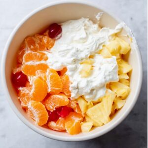 Ambrosia Fruit Salad Recipe 13 “Top-down food photography of a large white mixing bowl with ambrosia salad ingredients: whipped cream, mandarin orange segments, pineapple chunks, and maraschino cherries, natural soft light, clean marble background, minimal styling, high detail, realistic food texture, blog-style food photo”