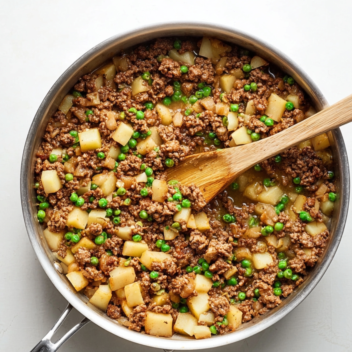 Easy baked beef empanadas | Golden & Freezer-Friendly 11 Ground beef empanada filling cooking in a stainless steel skillet with peas and diced potatoes, wooden spoon stirring, top-down view, clean white background, warm natural lighting, homemade comfort food, high-detail culinary photography