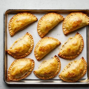 Easy baked beef empanadas | Golden & Freezer-Friendly 18 Freshly baked beef empanadas lined on a metal baking sheet with parchment paper, evenly spaced, golden brown crust, top-down flat lay composition, clean minimal background, bright natural light, professional food photography, high resolution, bakery style