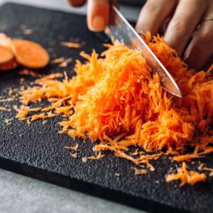 Easy Carrot Cupcake Recipe | Moist & Ready in 40 Minutes 15 Fresh carrots finely grated and chopped on a dark cutting board, hands holding a knife, close-up texture detail, bright contrast, clean kitchen prep scene.