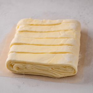 Chocolate Croissants Recipe 16 Butter block folded into croissant dough for lamination, visible layers of butter and dough, square dough shape, baking preparation process, neutral background, realistic food photography, high texture detail