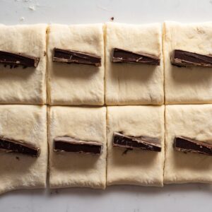 Chocolate Croissants Recipe 17 Raw croissant dough cut into even rectangles, dark chocolate batons placed on top of each piece, clean marble countertop, step-by-step baking process, natural soft light, minimal food styling, overhead shot