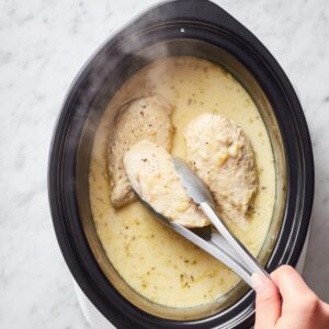Easy Crockpot Chicken Pot Pie 15 “Top-down food photography of a black slow cooker filled with creamy, light-yellow sauce and two tender chicken breasts partially submerged. A hand holding stainless steel kitchen tongs gently lifts one chicken breast, with steam rising to show warmth. The scene is set on a bright white marble countertop, natural soft lighting, realistic textures, cozy home-cooking vibe, high resolution, sharp focus.”