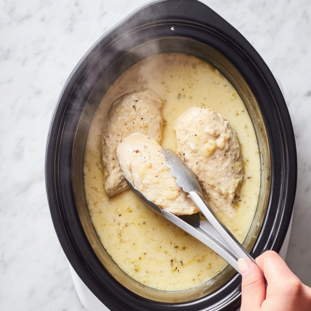 Easy Crockpot Chicken Pot Pie 11 “Top-down food photography of a black slow cooker filled with creamy, light-yellow sauce and two tender chicken breasts partially submerged. A hand holding stainless steel kitchen tongs gently lifts one chicken breast, with steam rising to show warmth. The scene is set on a bright white marble countertop, natural soft lighting, realistic textures, cozy home-cooking vibe, high resolution, sharp focus.”