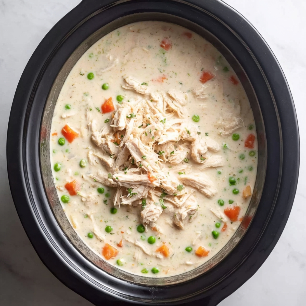 Easy Crockpot Chicken Pot Pie 12 “Top-down food photography of a slow cooker filled with creamy chicken soup, shredded chicken piled in the center, visible peas and diced carrots in a rich off-white broth, black slow cooker insert, minimal white marble background, soft natural lighting, high detail, realistic textures, cozy homemade comfort food, professional food photography, sharp focus, 4k”