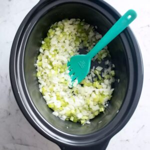 Easy Crockpot Chicken Pot Pie 14 “Top-down view of a black slow cooker pot containing finely chopped white onions and sliced celery being sautéed, with a teal silicone spatula resting inside. Minimalist kitchen scene, soft natural lighting, white marble countertop background, realistic food photography, high detail, neutral tones.”