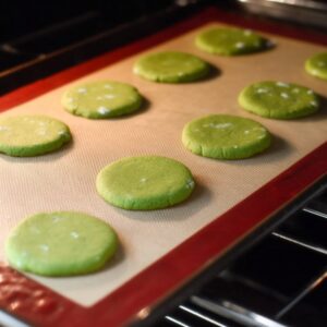 Easy Matcha Cookies Recipe 16 Overhead food photography of green matcha cookie dough rounds baking on a silicone baking mat inside an oven, evenly spaced cookies, soft oven light, baking process shot, realistic texture, high detail, home baking style