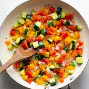 Best Omelette Recipe 14 “Overhead shot of sautéed diced vegetables including red and yellow bell peppers, zucchini, and onions in a white skillet, wooden spoon, glossy vegetables, fresh and colorful, natural daylight, healthy cooking style, high-resolution food photography