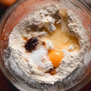 Best Red Velvet Cinnamon Rolls | Soft & Fluffy Recipe 15 Overhead view of cinnamon roll dough ingredients in a glass bowl, including flour, egg, sugar, butter, and vanilla.