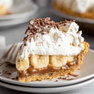 A close-up of a decadent slice of Banoffee Pie, topped with whipped cream, chocolate shavings, and a layer of banana slices on a buttery graham cracker crust.
